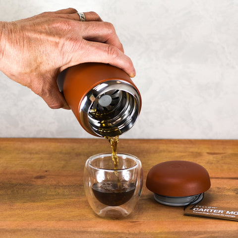 Fellow Carter Move travel mug being poured into a double-walled glass cup on a wood table, lid removed and set aside