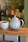 For Life Dew ceramic teapot in Natural Cotton with matching cup on a wooden surface, shown in tea shop setting