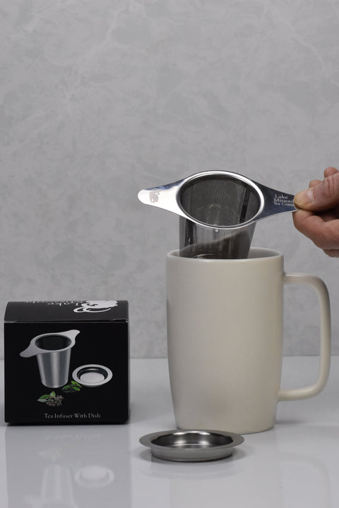 Hand lifting stainless steel loose leaf tea infuser from a cream ceramic mug, with metal drip dish and product box on white surface